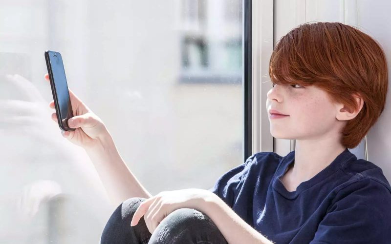 Rødhåret dreng sidder i vindueskam og kigger på en telefon.