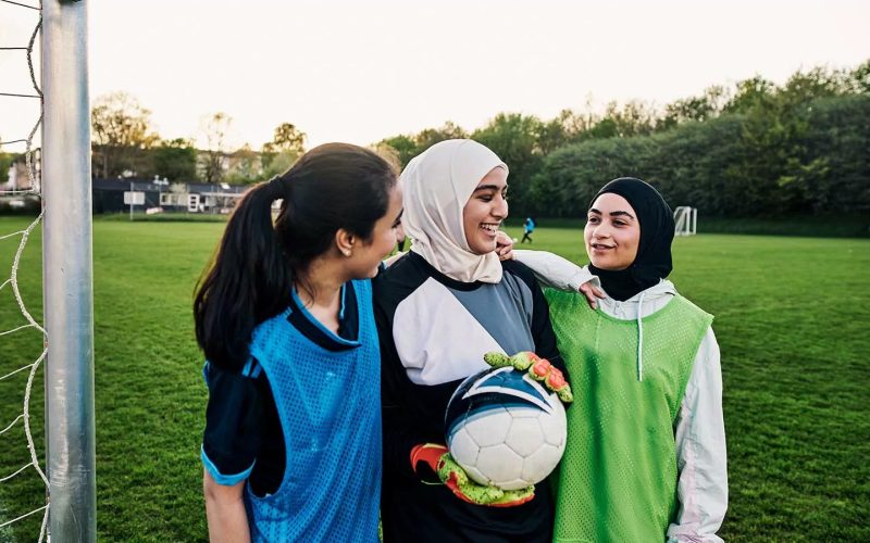 Tre piger står på en fodboldbane og smiler, den midterste med en fodbold i hånden.