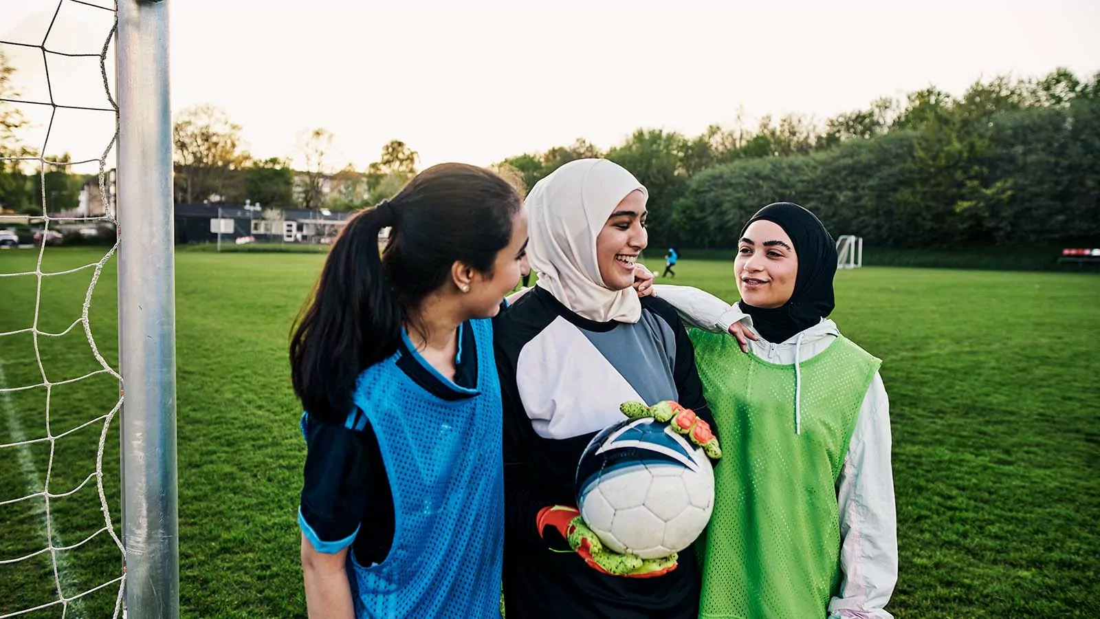 Tre piger står på en fodboldbane og smiler, den midterste med en fodbold i hånden.