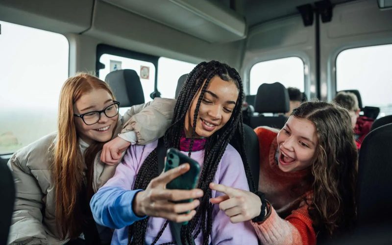 Tre piger sidder i en bus og kigger på en mobil